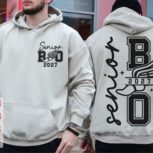 May include: A beige hooded sweatshirt featuring a black graphic print. The front displays the text Senior 2027 with a winged shoe icon. The back features the word Senior in script, the letter B, a winged shoe, the year 2027, and the letter O.