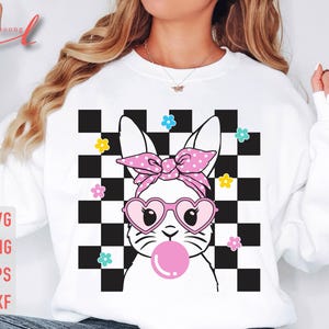 May include: White sweatshirt featuring a cartoon bunny with heart-shaped sunglasses and a pink bow, blowing a bubble. The bunny is set against a black and white checkered background with colorful flower accents. The image includes the text "SVG PNG EPS DXF".