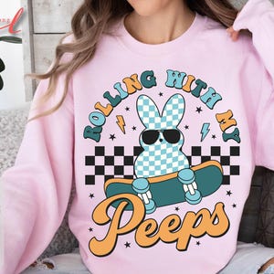 May include: A light pink sweatshirt with the text "Rolling with my Peeps." The graphic features a bunny wearing sunglasses on a skateboard, with a checkered background and lightning bolts.