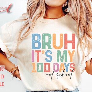 May include: A white t-shirt with the text "BRUH IT'S MY 100 DAYS -of school" in rainbow colours with a distressed look.
