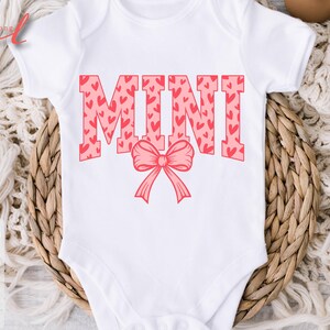 May include: White baby onesie with the word "MINI" in pink letters with a pink heart pattern. A pink bow is below the word.