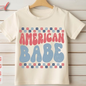May include: A white t-shirt with a red and blue design. The design features the text "AMERICAN BABE" in a retro font with stars and checkered patterns.