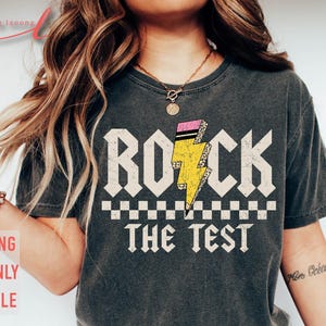 以下が含まれることがあります： 白い文字で「Rock the Test」と書かれた灰色のTシャツ。テキストの上に、ピンクと黒のアクセントが付いた黄色の稲妻があります。