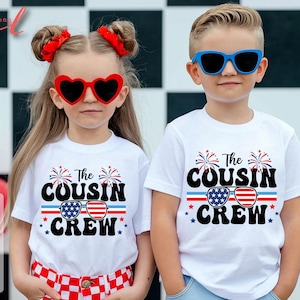 May include: Two children wearing white t-shirts with the text "The Cousin Crew" in black, red, and blue. The design includes sunglasses with an American flag pattern. One child wears heart-shaped sunglasses, the other wears blue sunglasses.