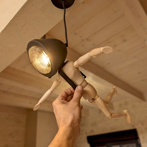 Może przedstawiać: Drewniany manekin z czarnym oświetleniem przymocowanym do głowy. Manekin jest zawieszony na suficie i znajduje się w pozycji lotu. Oświetlenie jest włączone i oświetla pomieszczenie.