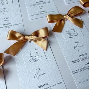 以下が含まれることがあります： 金色のサテンリボンが付いたエレガントな白いメニューカード。「The Menu」の文字がスタイリッシュな筆記体で書かれ、メニュー項目が下にリストされています。結婚式や特別なイベントに最適です。