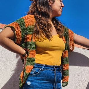 Könnte beinhalten: Eine Frau trägt eine grün-orange gestreifte Häkeljacke mit einem gelben Tanktop und blauen Jeans.