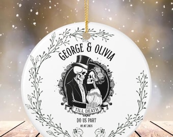 May include: A round white ceramic ornament featuring a black ink illustration of two skeletons in wedding attire within an ornate frame. The text reads GEORGE & OLIVIA, TILL DEATH DO US PART, and the date 09.07.2026. A gold string is attached to the top.