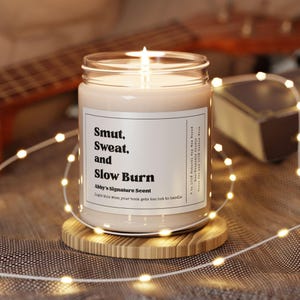 May include: A scented candle in a glass jar with the text "Smut, Sweat, and Slow Burn" on a white label. The candle is on a wooden coaster, with fairy lights around it. A guitar and a book are in the background.