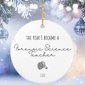 Può includere: Ornamento natalizio rotondo in ceramica bianca con cordino dorato. Il testo recita "THE YEAR I BECAME A Forensic Science Teacher" in corsivo nero. Sotto c'è una grafica di impronta digitale e lente d'ingrandimento, con "20XX" in basso.