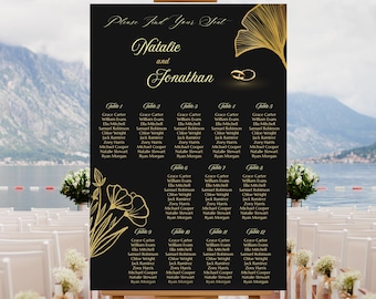May include: A black wedding seating chart on an easel. The chart features gold floral accents, the names "Natalie and Jonathan," and guest seating assignments. The background shows a scenic view of water and mountains.