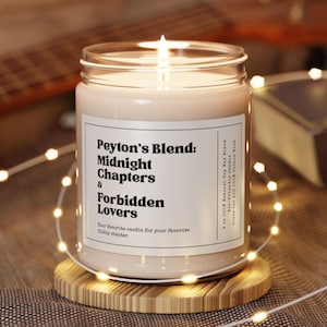 Op de afbeelding: Een brandende kaars in een glazen pot op een houten onderzetter. De kaars heeft een label met de tekst "Peyton's Blend: Midnight Chapters & Forbidden Lovers". De kaars is omgeven door een lichtsnoer.