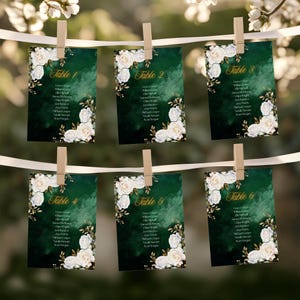 May include: Six emerald green table cards with white floral and gold leaf accents are clipped to a white ribbon. Each card has the word "Table" followed by a number and a list of names. The cards are decorated with white roses.