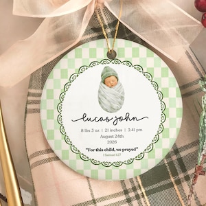 Op de afbeelding: Een rond keramieken ornament met een lichtgroen en wit ruitpatroon, voorzien van een centrale illustratie van een ingebakerde baby. De tekst bevat de naam Lucas John, geboortegegevens van 8 lbs 3 oz, 21 inches, 3:41 pm, 24 augustus 2026 en het citaat For this child, we prayed 1 Samuel 1:27.