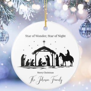 May include: A white ceramic Christmas ornament with a gold string. The ornament features a black silhouette of the Nativity scene, with text that reads "Star of Wonder, Star of Night", "Merry Christmas", and a family name.
