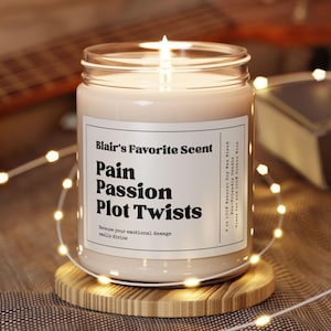 以下が含まれることがあります： 「Blair's Favorite Scent: Pain, Passion, Plot Twists」と書かれた、ガラス瓶に入った、火の灯った大豆ワックスブレンドのキャンドル。木製のコースターの上に置かれ、ストリングライトで囲まれています。「Because your emotional damage smells divine」というテキストもラベルにあります。