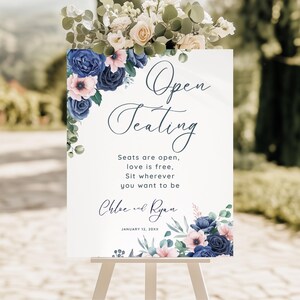 May include: A white sign with the words "Open Seating" in elegant script, adorned with blue and pink floral arrangements. The sign includes the text "Seats are open, love is free, Sit wherever you want to be" and the names "Chloe and Ryan".