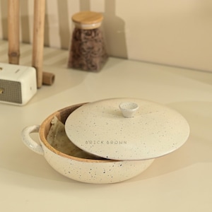 May include: A speckled off-white ceramic bowl with a matching lid. The lid has a small knob and the words "BRICK BROWN" printed on it. The bowl has a wooden interior and a handle. The bowl contains a folded piece of food.