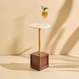 Könnte beinhalten: Ein moderner Beistelltisch mit einer weißen, kreisförmigen Tischplatte, getragen von einer goldfarbenen Stange und einem dunkelbraunen Holzsockel. Ein Glas bernsteinfarbener Flüssigkeit mit grünen Blättern steht auf dem Tisch. Für den Innenbereich.