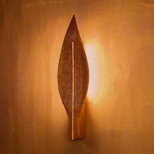 Op de afbeelding: Een houten wandlamp in de vorm van een blad, met een verticale lichtstrip in het midden. De lamp is gemaakt van donkerbruin hout en is bevestigd aan een muur met een warme, gouden tint. De lichtstrip geeft een zachte gloed.