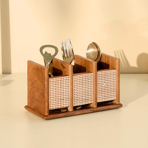 Könnte beinhalten: Ein hölzerner Utensilienhalter mit drei Fächern, jedes mit einem gewebten Rattanpaneel. Der Halter enthält einen Flaschenöffner, eine Gabel und einen Löffel. Das Holz hat einen warmen, rötlich-braunen Ton, und das Design ist minimalistisch und funktional.