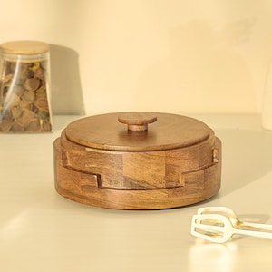 May include: A round, wooden container with a lid and a small handle. The container is a warm brown color and has a geometric design. The text "BRICK BROWN" is visible on the side. A glass jar with a wooden lid and a pair of tongs are also in the image.