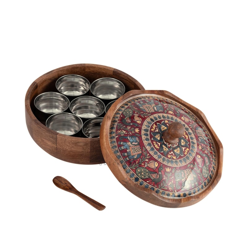 May include: A wooden spice box with a decorative lid featuring a detailed, colorful pattern. The box contains several small stainless steel containers. A small wooden spoon rests beside the open box, suggesting its use for measuring spices.