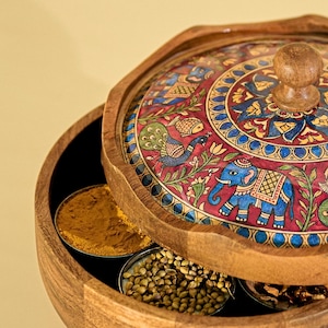 May include: Wooden spice box with a decorative lid featuring a painted design of an elephant, peacock, and floral patterns in red, blue, and gold. The box contains small metal containers filled with spices.