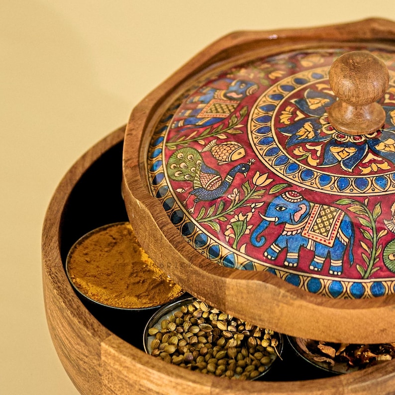 May include: Wooden spice box with a decorative lid featuring a painted design of an elephant, peacock, and floral patterns in red, blue, and gold. The box contains small metal containers filled with spices.