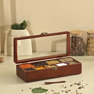 May include: A dark wooden spice box with a glass lid, containing eight compartments of different spices. A small wooden spoon lies beside the box. Two glass jars with wooden lids are visible in the background.