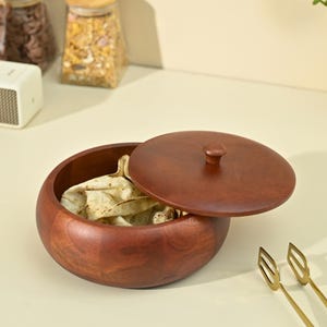 May include: A dark brown wooden bowl with a lid, containing flatbread. The bowl is round with a smooth finish, and the lid has a small knob. Gold-coloured serving tongs are visible in the foreground, suggesting a serving dish.