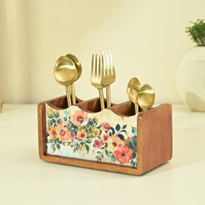 Può includere: Un portaposate in legno con tre scomparti, caratterizzato da un design floreale con fiori rosa, arancioni e gialli. All'interno sono posizionati cucchiai e forchette color oro, creando un accessorio da cucina decorativo e funzionale.