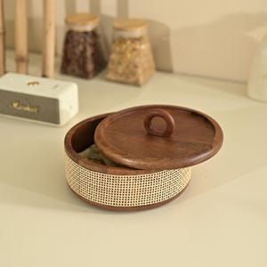 May include: A round wooden storage box with a woven rattan base and a wooden lid. The lid has a circular handle. The box is partially open, revealing the contents inside. The box is on a light-colored surface.