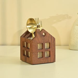 Op de afbeelding: Een huisvormige houten bestekhouder met een donkerbruine afwerking en lichtgekleurde gevlochten rieten panelen in de vensteropeningen. De houder bevat een goudkleurige metalen lepel en vork. Aan de voorzijde bevindt zich een kleine ronde uitsparing.