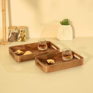 May include: Two rectangular wooden serving trays with vertical ribbed detailing on the exterior sides and integrated handles. Each tray contains a clear glass tumbler filled with amber liquid and a small pastry item placed on the smooth interior base.