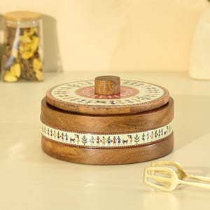 May include: A round wooden container with a lid. The lid features a circular design with red, beige, and brown patterns. The container's body has a decorative band with figures. A jar with a wooden lid is in the background.