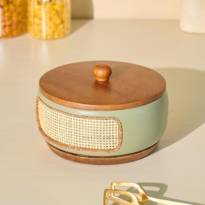 May include: A round, lidded container with a light green body, a woven rattan panel, and a wooden lid. The lid has a small, rounded wooden knob. The container sits on a wooden base, suggesting a decorative storage item.