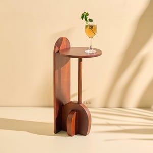 May include: A contemporary side table crafted from dark wood, featuring a circular top and a distinctive curved design. A glass of amber liquid with a sprig of greenery rests on the tabletop. The table is a stylish accent piece.