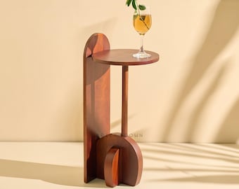 Puede incluir: Una mesa auxiliar moderna de madera oscura con una parte superior circular y un diseño curvo y arquitectónico. Un vaso de líquido ámbar con una ramita de vegetación descansa sobre la mesa. La mesa es una pieza de acento elegante.