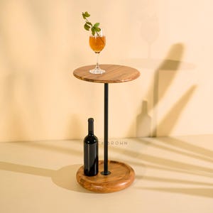 Puede incluir: Una mesa auxiliar de madera con tapa y base redondas, conectadas por un poste de metal negro. Una botella de vino y una copa de líquido ámbar con una ramita de vegetación están sobre la mesa. La mesa está sobre una superficie beige claro.