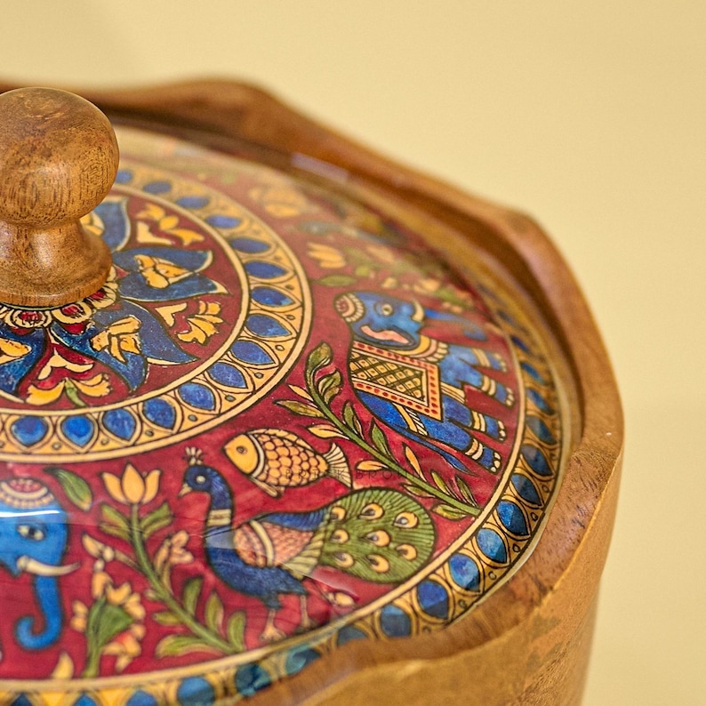 May include: A wooden serving bowl with a decorative lid. The lid features a vibrant, hand-painted design with a red background, blue and yellow accents, and images of elephants, peacocks, and fish. The bowl has a wooden knob and rim.