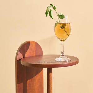 May include: A small, round, dark wood side table with a single stem glass of amber liquid and a sprig of greenery. The table has a curved back and a single leg. The text "BRICK BROWN" is printed on the table's surface.