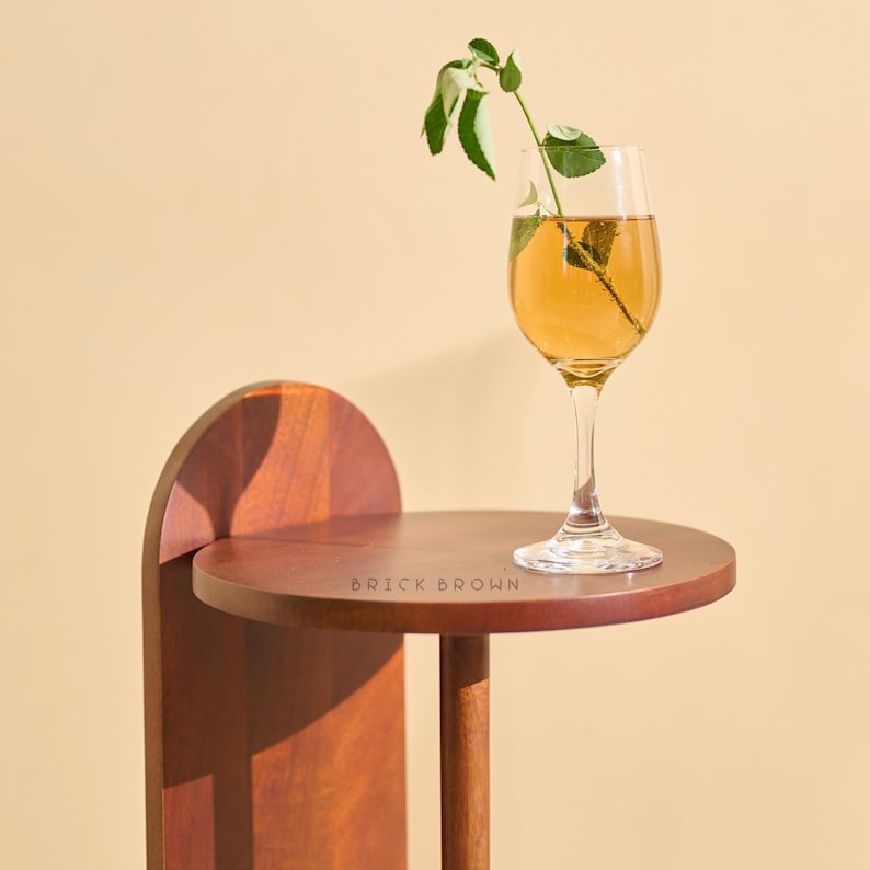 May include: A small, round, dark wood side table with a single stem glass of amber liquid and a sprig of greenery. The table has a curved back and a single leg. The text "BRICK BROWN" is printed on the table's surface.