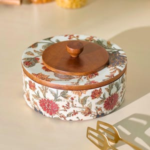 May include: A round, floral-patterned serving bowl with a wooden lid and knob. The bowl features a white background with red, orange, and green floral designs. The lid and base are made of wood. Gold serving utensils are in the foreground.