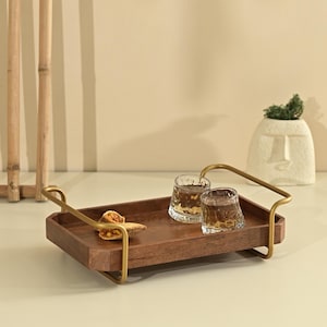 Könnte beinhalten: Ein rechteckiges Serviertablett aus Holz mit dunkelbraunem Finish und goldfarbenen Metallgriffen an beiden Seiten. Auf dem Tablett stehen zwei klare, strukturierte Glasbecher und ein kleines Stück Essen. Das Design hat klare Linien.