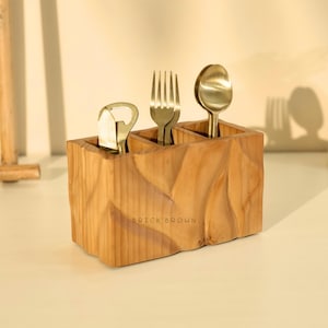 May include: A wooden utensil holder with three compartments, holding a bottle opener, two forks, and a spoon. The holder is light brown with a textured surface and the text "BRICK BROWN" on the front. The utensils are gold-colored.