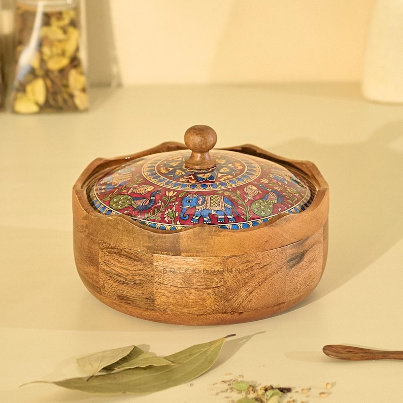 May include: A wooden bowl with a decorative lid. The lid features a colorful, intricate design with elephants and other patterns. The bowl is made of light brown wood and has a round shape.