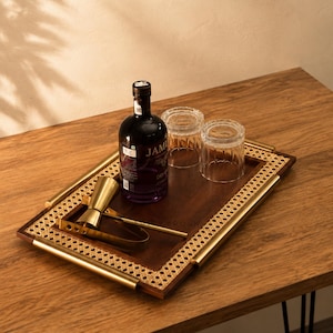 Puede incluir: Una bandeja rectangular con base de madera oscura, borde de rejilla tejida y asas de metal dorado pulido. La bandeja contiene una botella de vidrio oscuro, dos vasos estriados transparentes, un medidor y una cuchara de bar dorados.