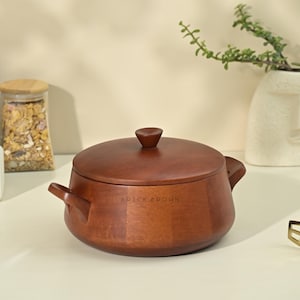 Puede incluir: Una olla de cocina de madera con tapa y asas laterales. La olla es de color marrón oscuro y tiene las palabras "BRICK BROWN" impresas en el lateral. Un tarro de cristal con tapa de madera y un jarrón blanco están en el fondo.