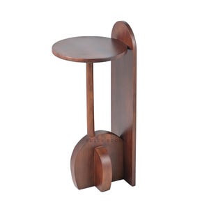 May include: A dark brown wooden side table with a round top, supported by a slender stem and a geometric base. The table has a tall, arched back panel. The brand name "BRICK BROWN" is visible on the base.